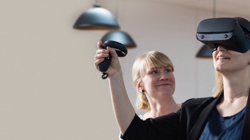 En ung dame bruker VR-briller. En annen dame står litt i bakgrunnen og følger smilende med. 