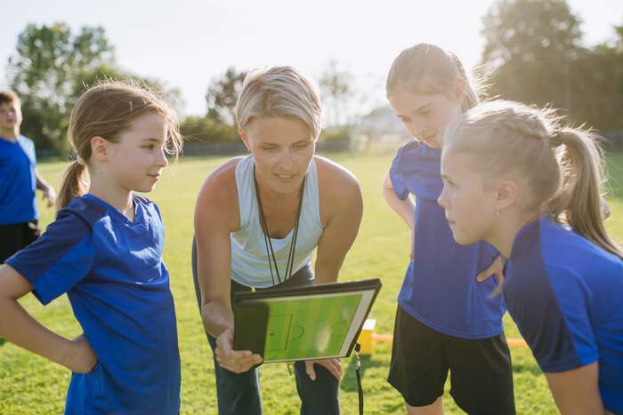 Illustrative image of a coach standing with a group of girls’ footballers on a football pitch
