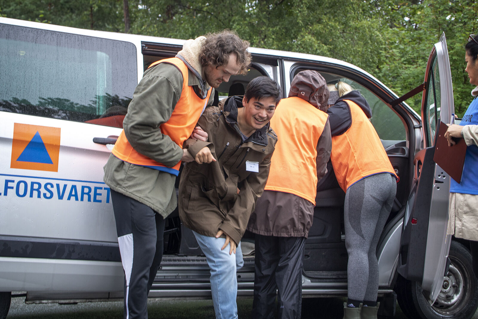 Bildet viser tre personer i oransje refleksvester ved en bil. En av personene med oransje vest hjelper en som spiller skadet ut av bilen.