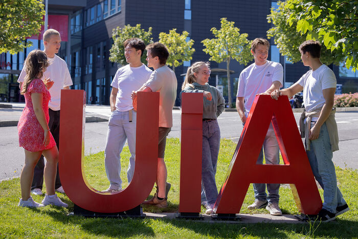 Røde UiA bokstaver utenfor Campus Grimstad, omgitt av smilende studenter i fint vær. Sommer.