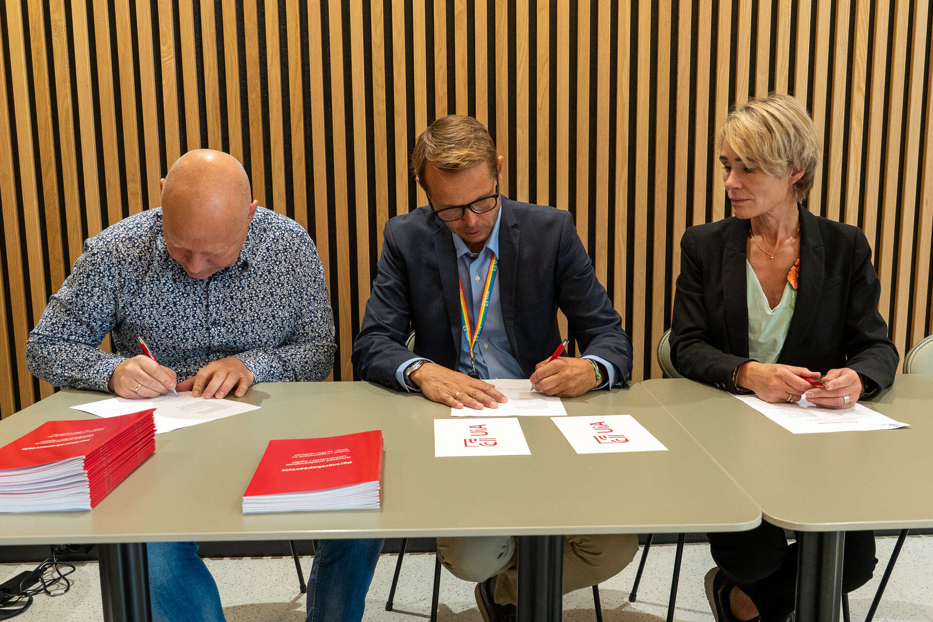 Bildet viser tre personar som sit bak eit bord, signerer