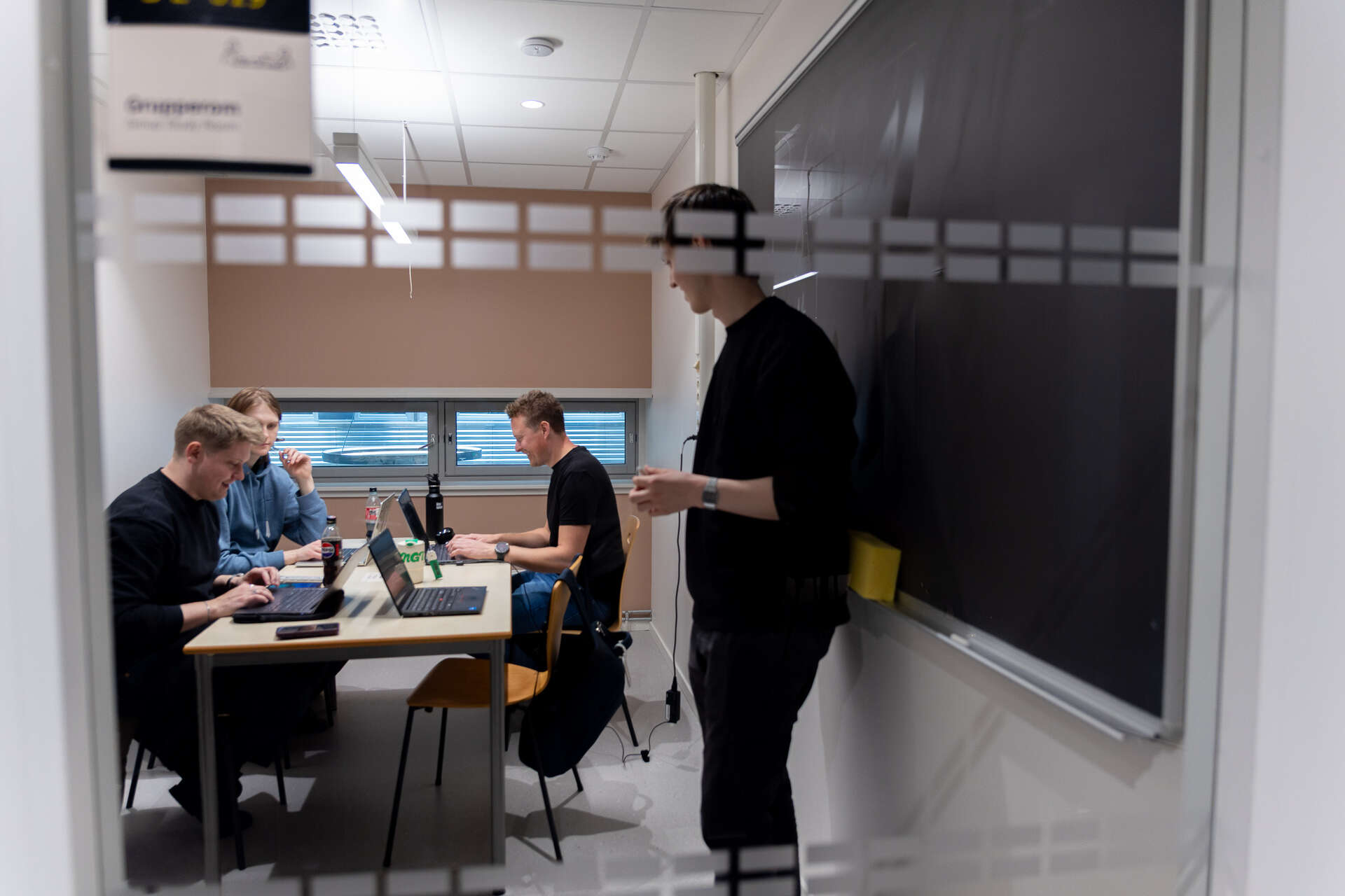 Sett gjennom glassveggen: Einar Alme står ved tavlen mens tre medstudenter i 1000BYTE sitter rundt et bord med bærbare datamaskiner.