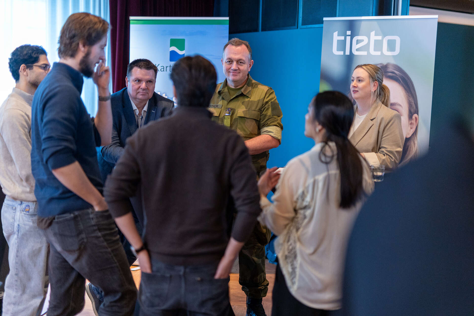 Studenter står rundt et samtalebord foran bannere fra Kartverket og Tieto. En representant i militær uniform og to andre representanter står på motsatt side av bordet.