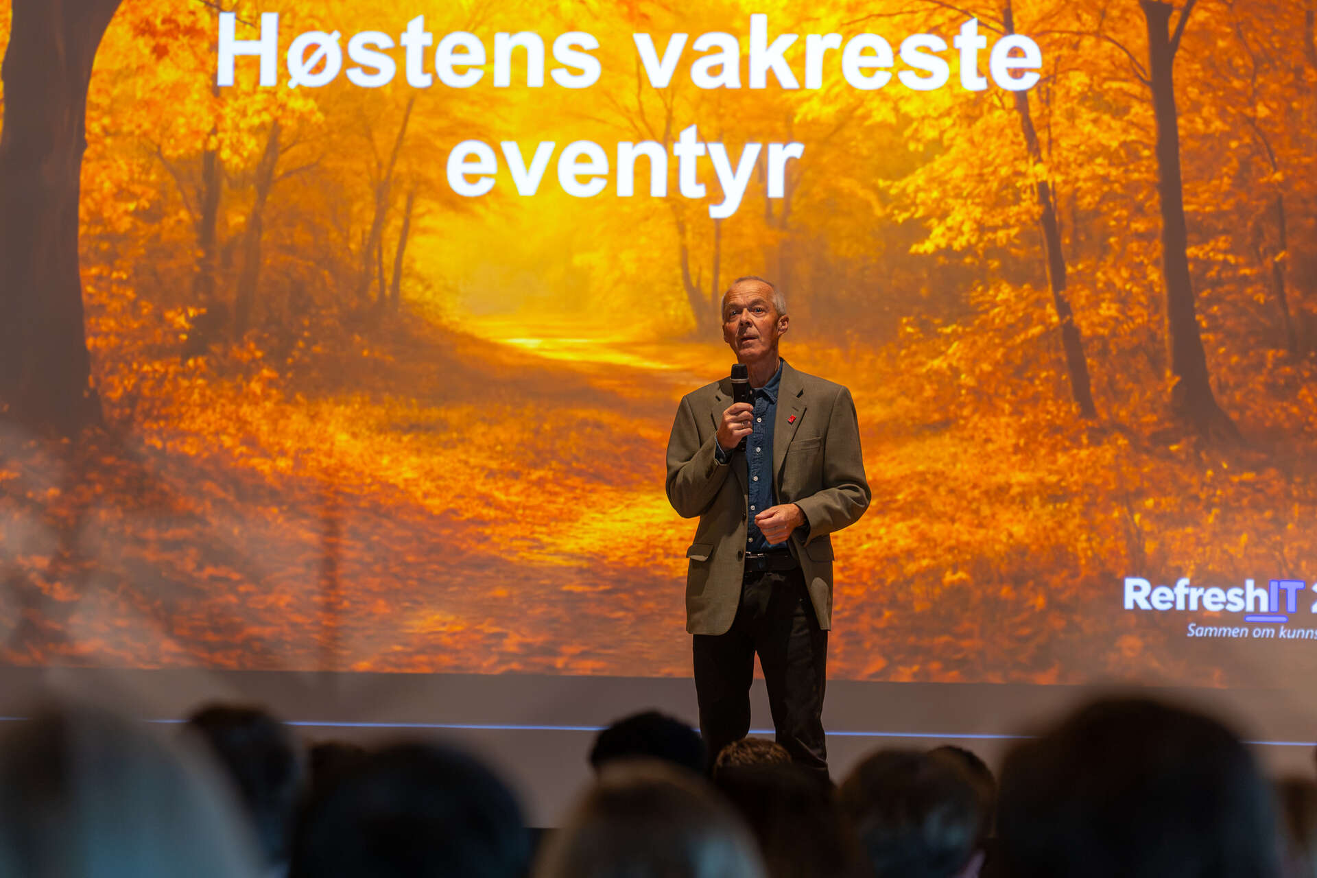 Hallgeir Nilsen med mikrofon på scenen foran en høstfarget RefreshIT-presentasjon. Publikum sitter i forgrunnen.