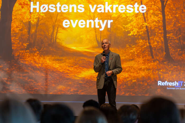 Hallgeir Nilsen med mikrofon på scenen foran en høstfarget RefreshIT-presentasjon. Publikum sitter i forgrunnen.