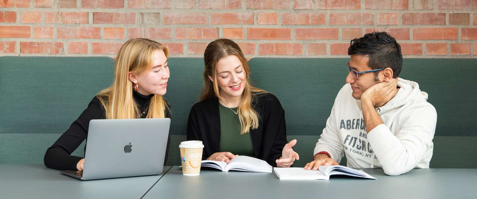 Three students sitting in a sofa in the faculty area, working with textbooks and computer, cup of coffee