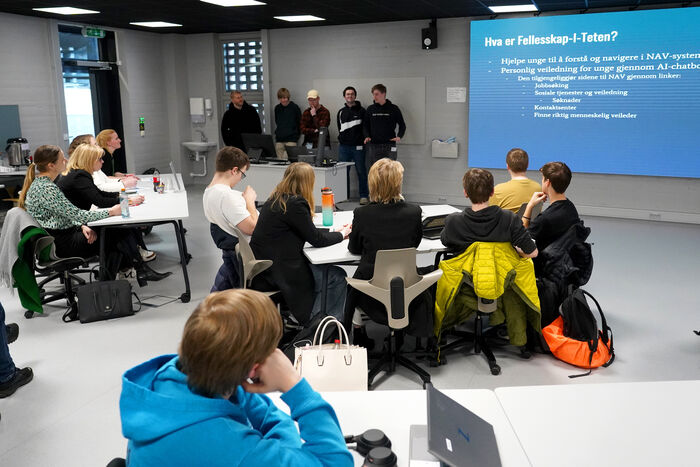 Fem unge studenter holder en presentasjon på en skjerm foran et dommerpanel og andre deltakere. 
