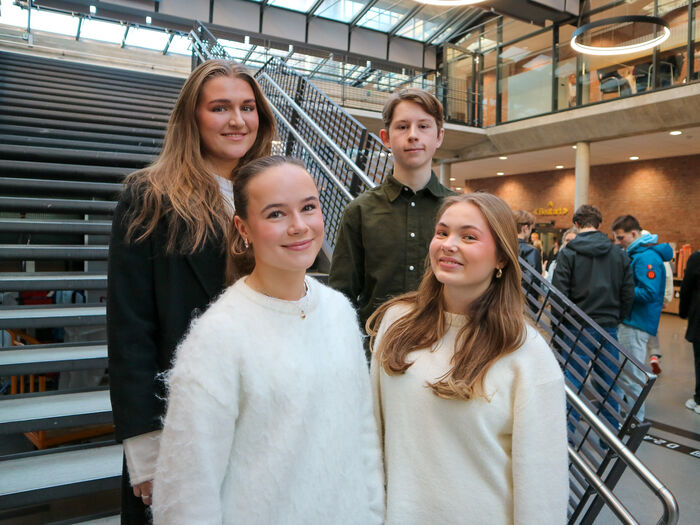 Tre jente og en gutt står sammen og smiler, studenter i bakgrunnen.