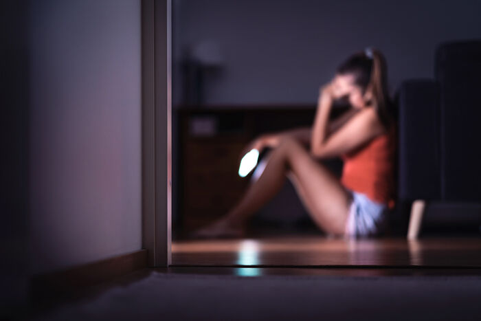 Bildet viser et uskarpt bilde av en dame som lener hodet i hånda og holder en telefon som lyser i den andre