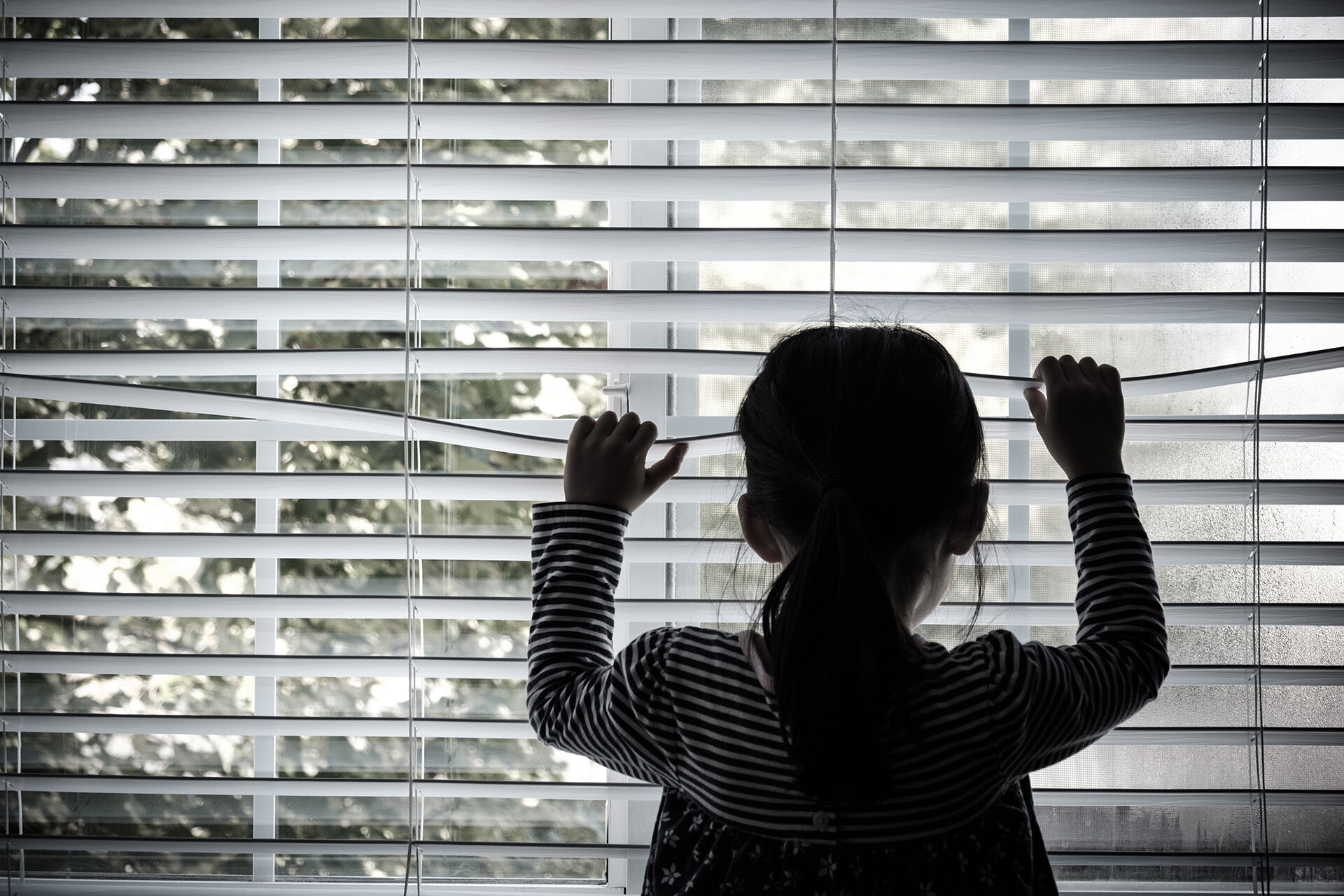 Bildet viser et barn som står ved et vindu og titter gjennom persiennene