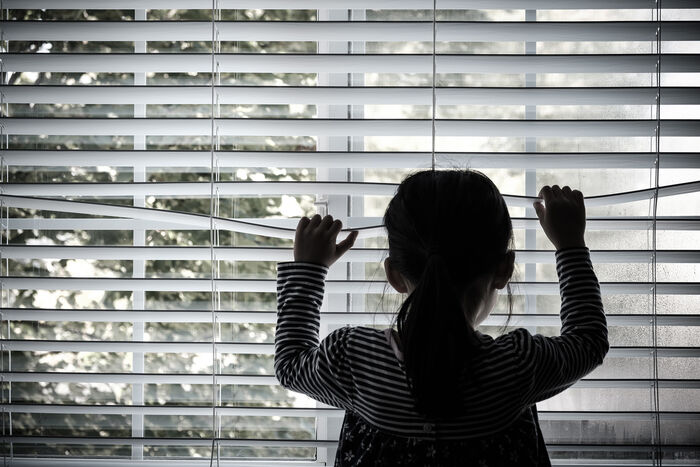 Bildet viser et barn som står ved et vindu og titter gjennom persiennene