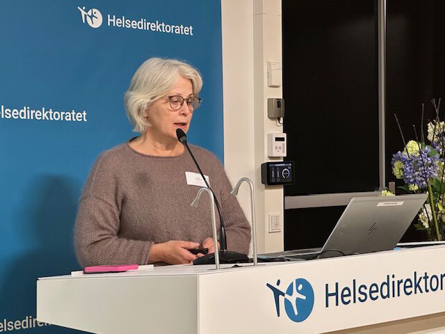 kvinne med grått hår og briller som står foran en mikrofon og presenerer, brun genser