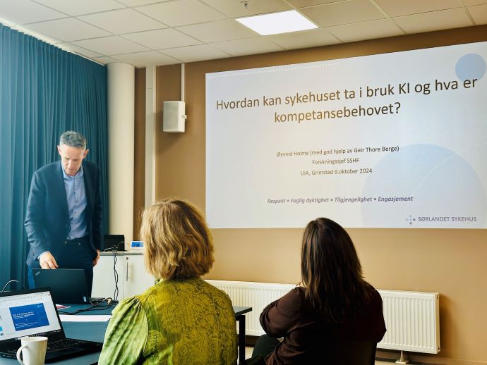 mann i sort jakke og blå skjorte stående ved et bord, klar til å presentere