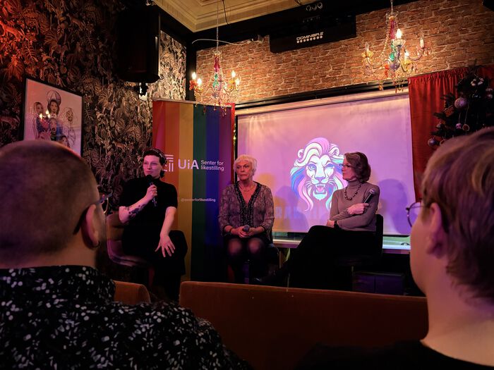 Mary Ann Skretteberg Warenius, Esben Esther Pirelli Benestad og Elsa Mari Almås sit på ei lita scene på Barrikaden. Mary Ann introduserer Esben Esther og Elsa før filmframsyninga og held ein mikrofon. Alle tre sit framfor eit prosjektorlerret med Barrikadens løve-logo i regnbogefargar. Til venstre står ein regnbogefarga rollup frå UiA – Senter for likestilling. I forgrunnen ser ein to publikumsdeltakarar vende mot scena. Det er stemningsfull, dempa belysning frå to regnbogefarga lysekroner som heng frå taket.