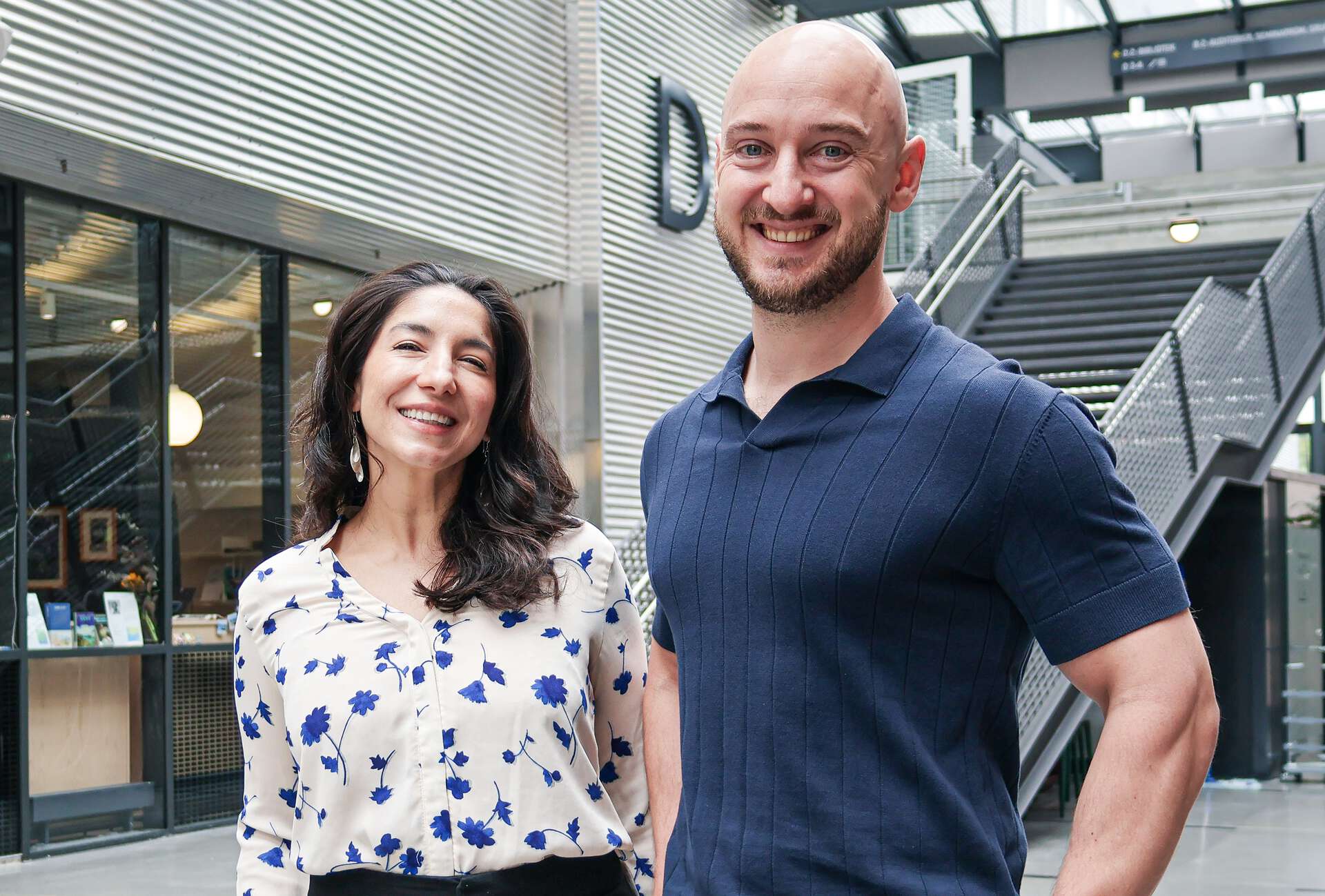 On the left, Adriana is standing, smiling, next to a smiling Lee; they are both in the Vrimlehallen on the Kristiansand Campus
