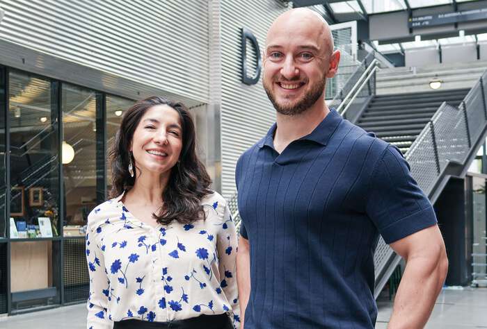 On the left, Adriana is standing, smiling, next to a smiling Lee; they are both in the Vrimlehallen on the Kristiansand Campus