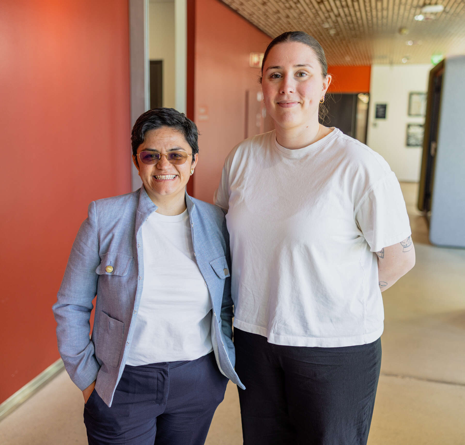 Cinthya smiler med tennene og har på seg en blå blazer, Thea smiler inn i kamera og har på seg en hvit t-skjorte, de står i en rød gang