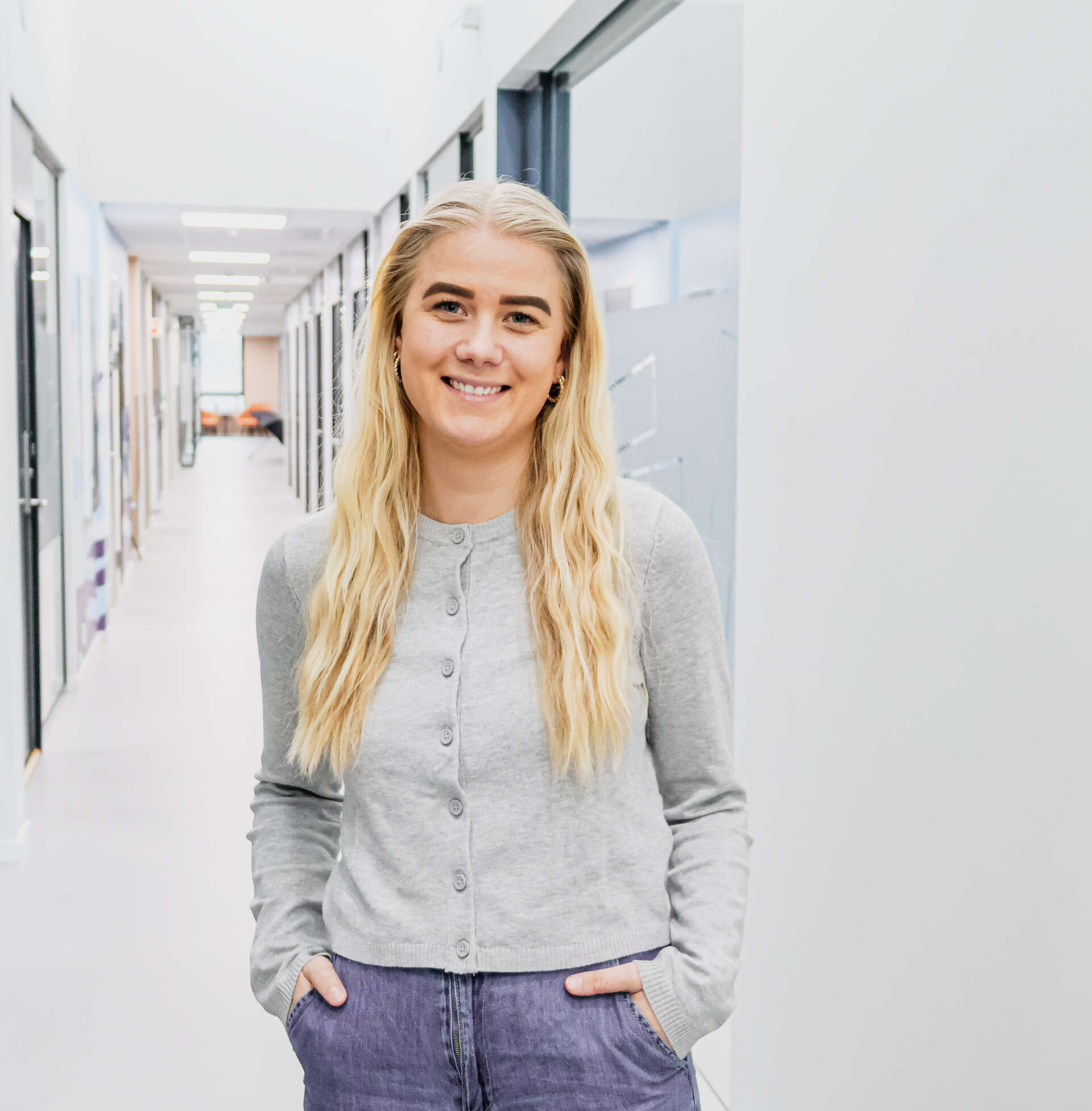 smilende kvinne med blondt hår, grå genser og olabukse, som står i en lys gang