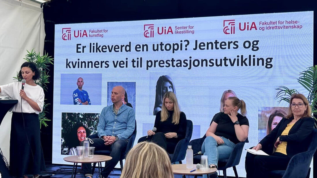 Bildet viser en paneldiskusjon på en scene. Det er fire personer som sitter i en sofa og en person som står ved et podium. Bak dem er det en stor skjerm med teksten "Er likeverd en utopi? Jenters og kvinners vei til prestasjonsutvikling". Det er også logoer for Universitetet i Agder (UiA) og Fakultet for helse- og idrettsvitenskap, samt Senter for likestilling.
