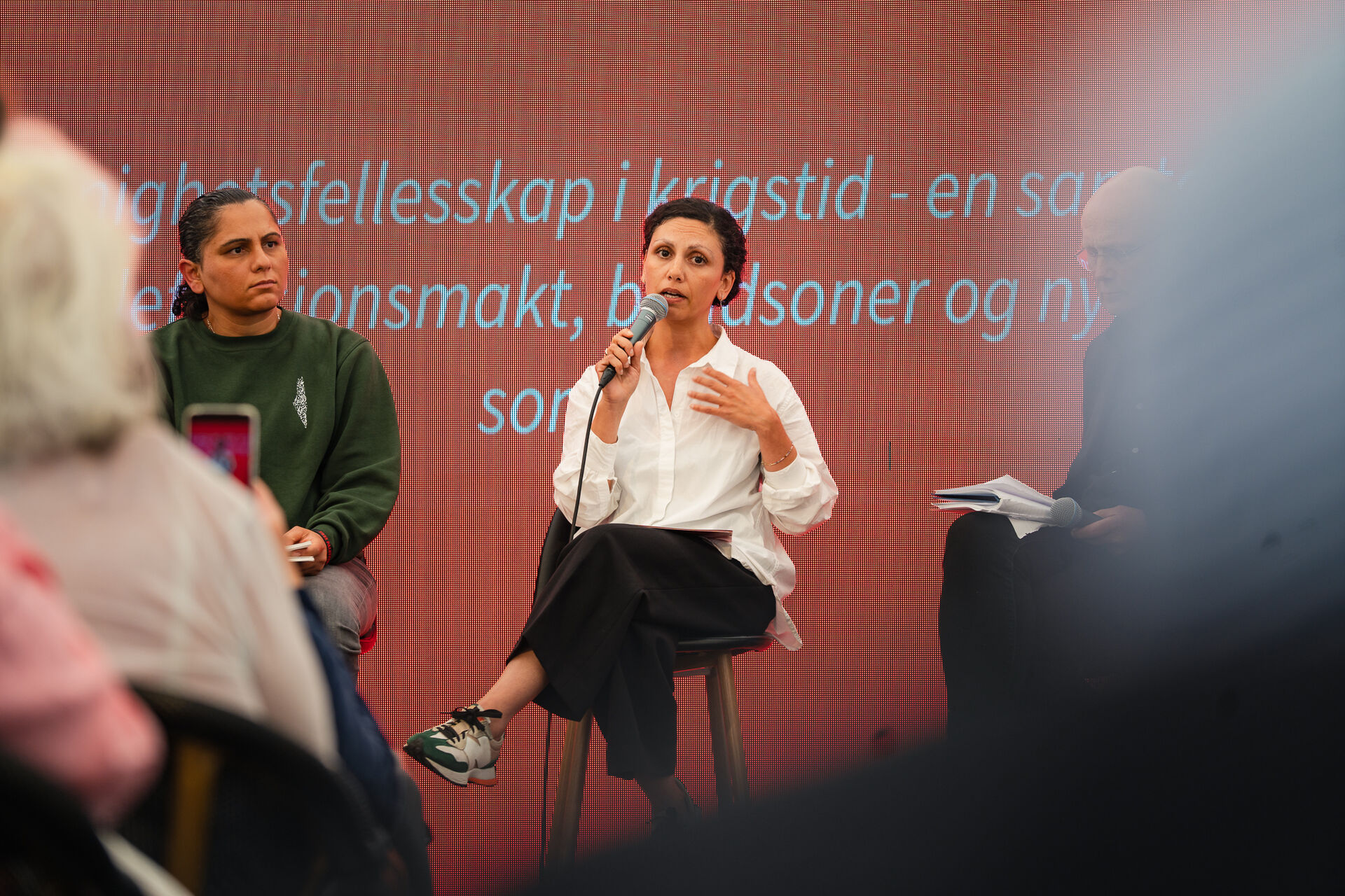 Bildet viser to personer samt en person som vare er delvis synlig. De sitter på en scene under en paneldiskusjon. Rasha Abdallah til høyre holder en mikrofon og snakker, mens personen til venstre lytter. Bak dem er det en stor skjerm med teksten: Uenighetsfelleskap i krigstid. Personen til høyre har på seg en hvit skjorte og mørke bukser, mens personen til venstre har på seg en mørk genser.
