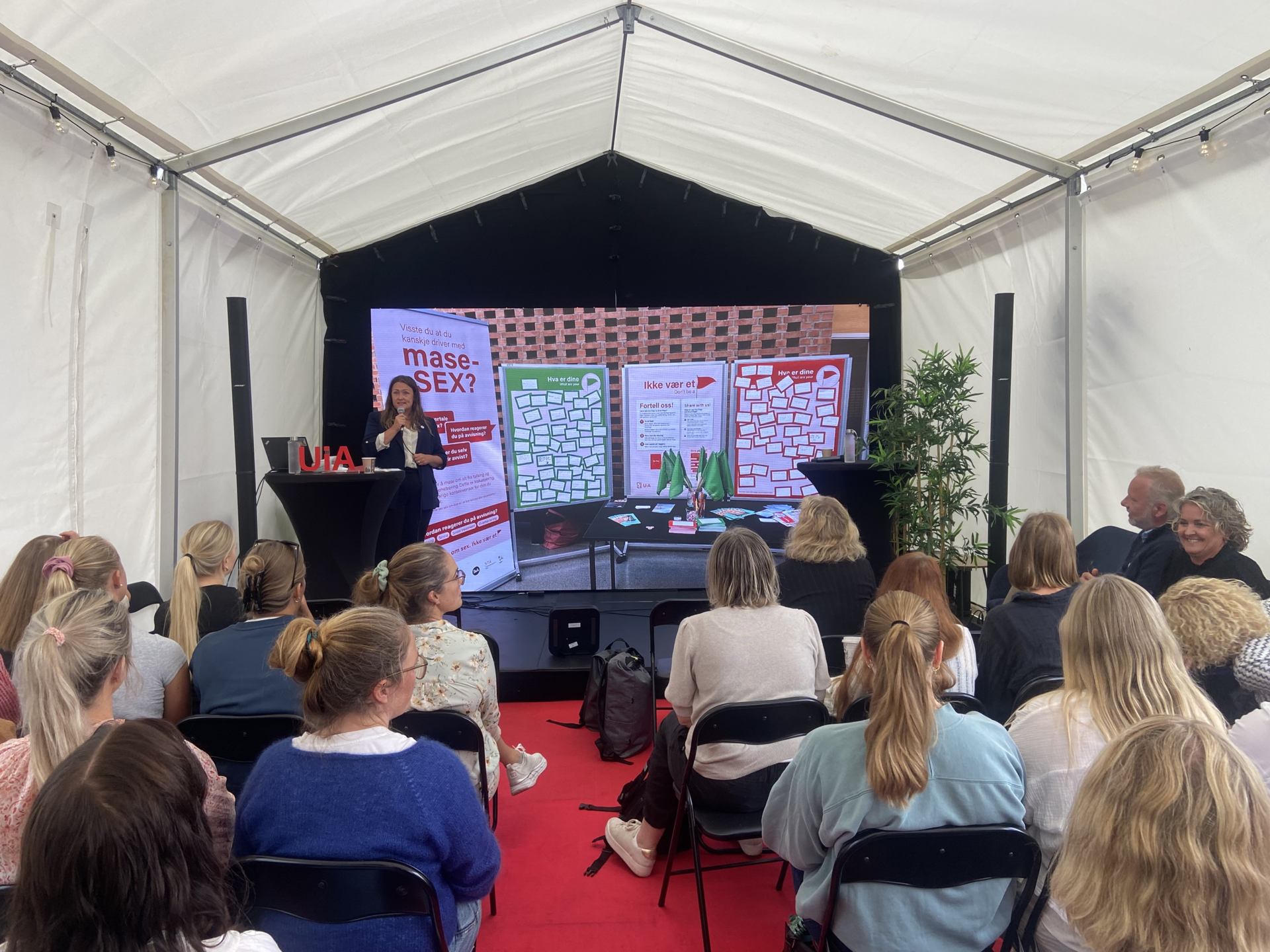Bildet viser Claudia på et podium der hun snakker i en mikrofon. Det sitter flere publikummere og lytter og ser på plakater som viser eksempler på hvordan kampanjen er laget.