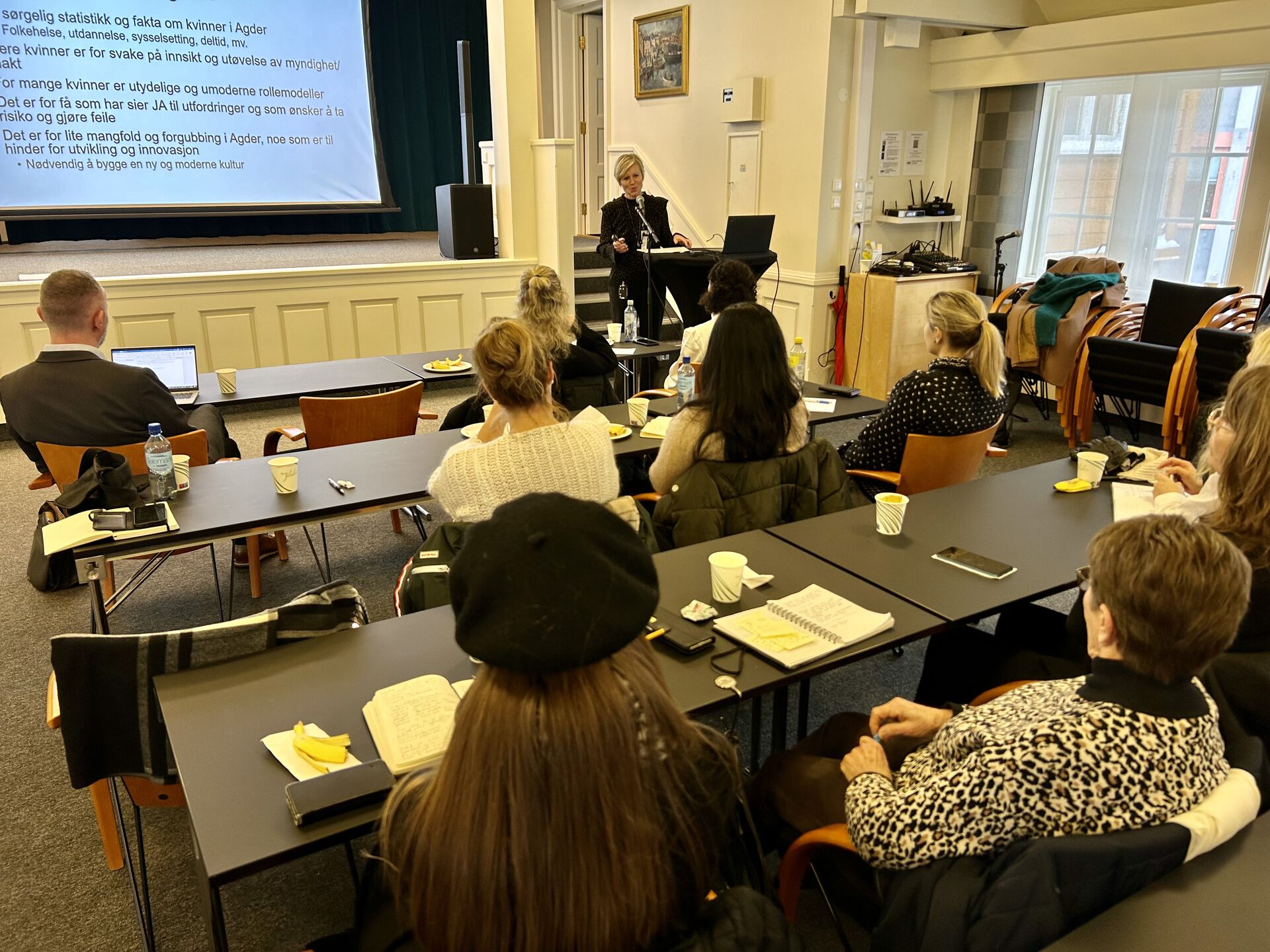 Bildet viser et foredrag i et innendørs miljø. Inger Hegna står foran og snakker, med en projektor bak som viser en presentasjon med tekst. Publikummet, som ser ut til å være hovedsakelig kvinner, sitter vendt mot foredragsholderen. De fleste har notatblokker og penner, og noen har kaffekopper. Rommet ser ut til å være en konferansesal eller et møterom med kunst på veggene. Det er også synlig en del av en dataskjerm i forgrunnen nederst på bildet.