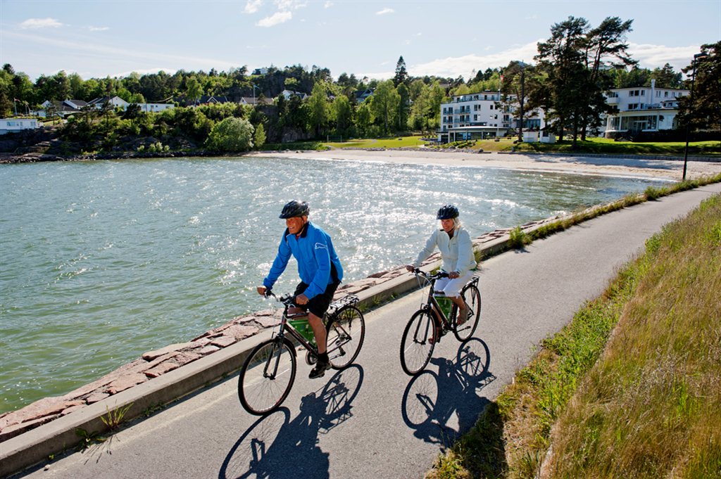 En mann og en kvinne sykler langs vannet en strålende solskinnsdag