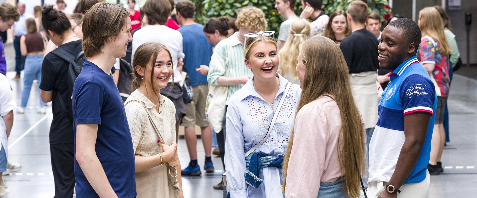 Fem studenter i vrimlehallen på UiA, med flere studenter i bakgrunnen.
