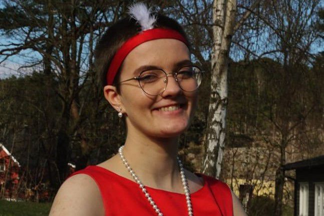 Victoria Abelsen poserer smilende i vårsola med koruniformen sin på. Det er en rød kjole med perlesmykke, og rødt hårbånd med en hvit fjær i på hodet.