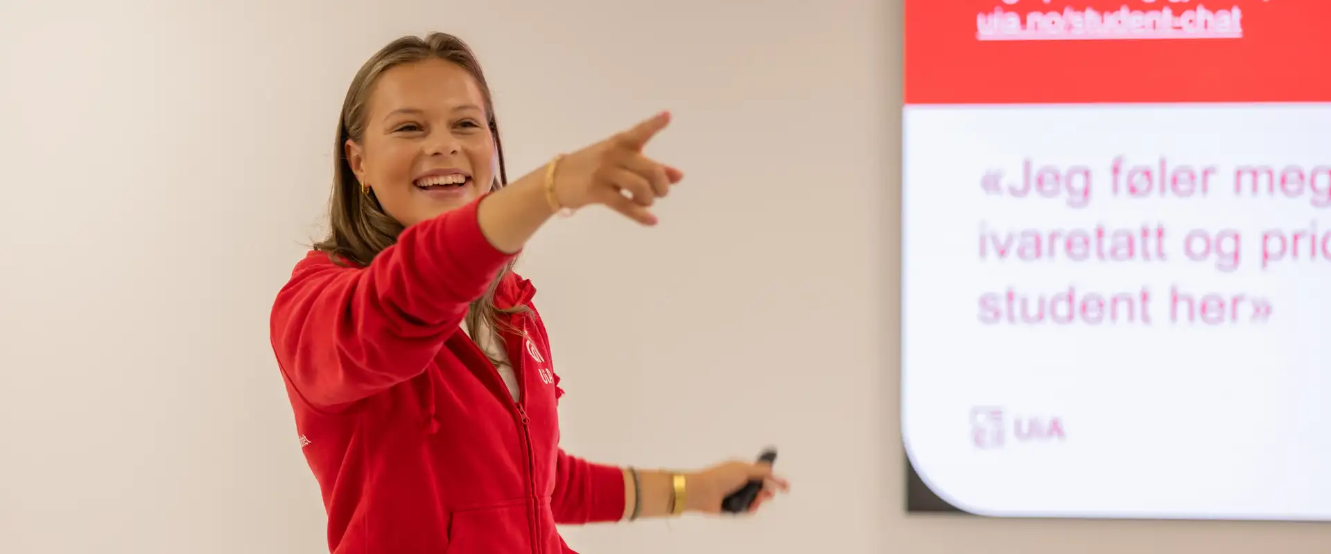 Kvinnelig studentambassadør står ved skjerm i klasserom og holder UiA-presentasjon