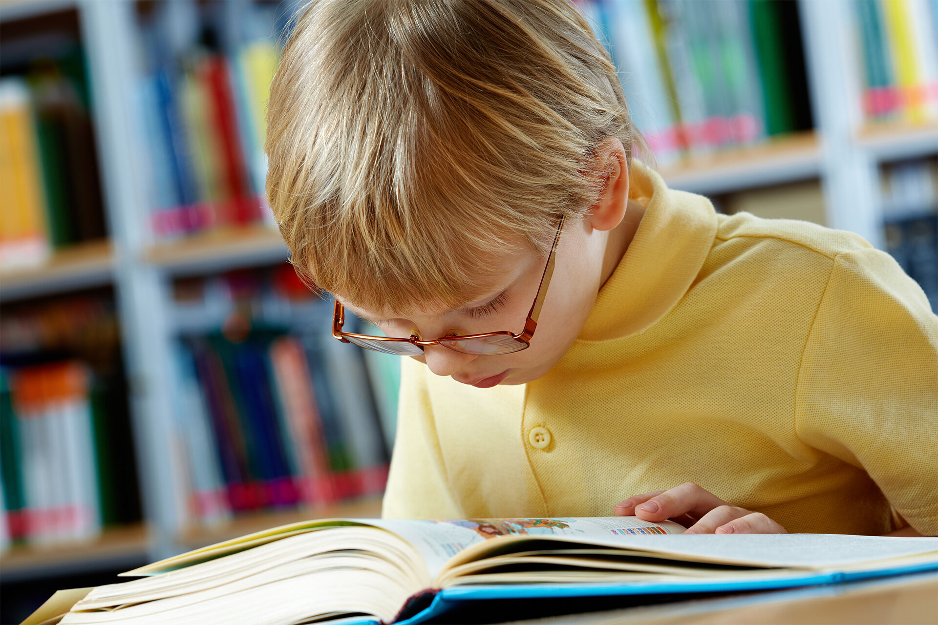 Gutt som sitter og leser i en bok på biblioteket. Han har på seg gul genser med krage, har blondt hår og briller.