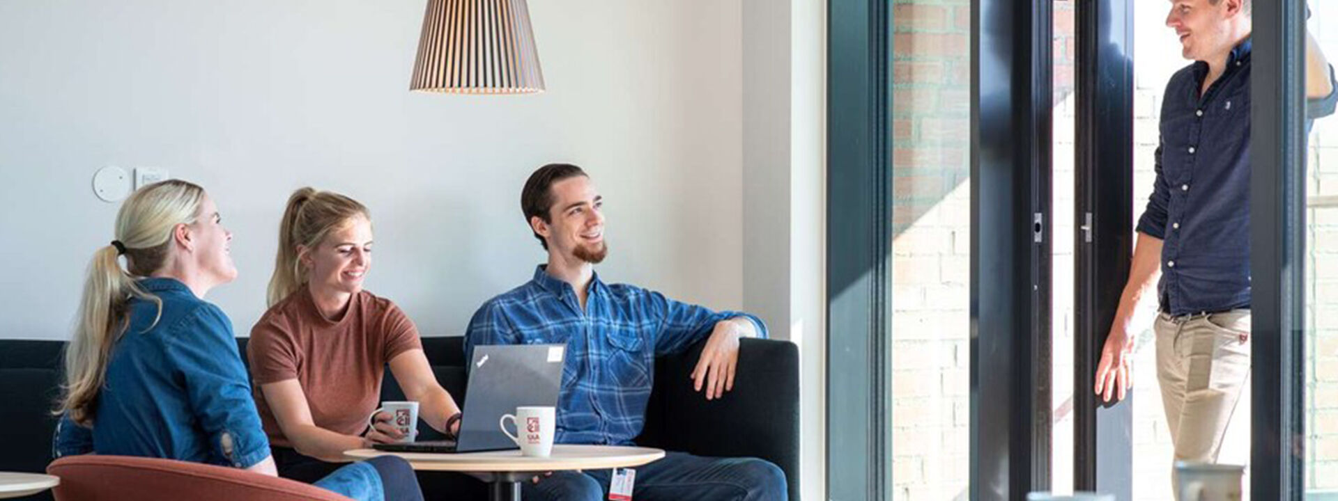 To kvinner og en mann sitter i en sofa, den ene kvinner arbeider på en PC. En fjerde mann står i en døråpning og tilsynelatende diskuterer noe med personene i sofaen.