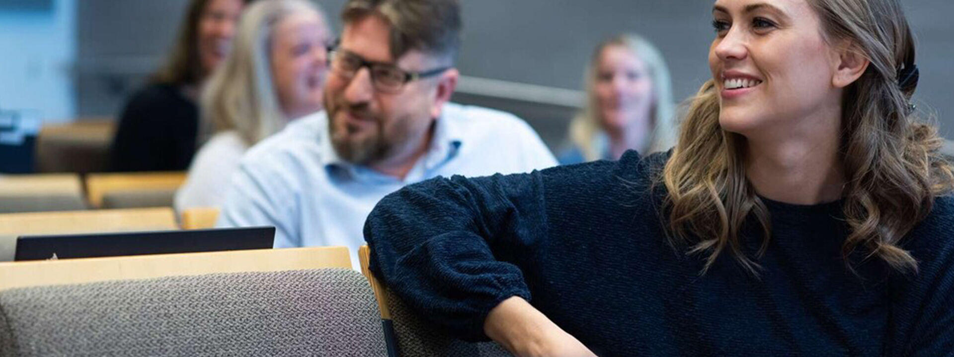 En mann i femtiårsalderen og en kvinne i trettiårene sitter i en forelesningssal. Kvinnen har armen over setet ved siden av seg og smiler, mannen sitter i bakgrunnen og smiler mot en pc-skjerm. 