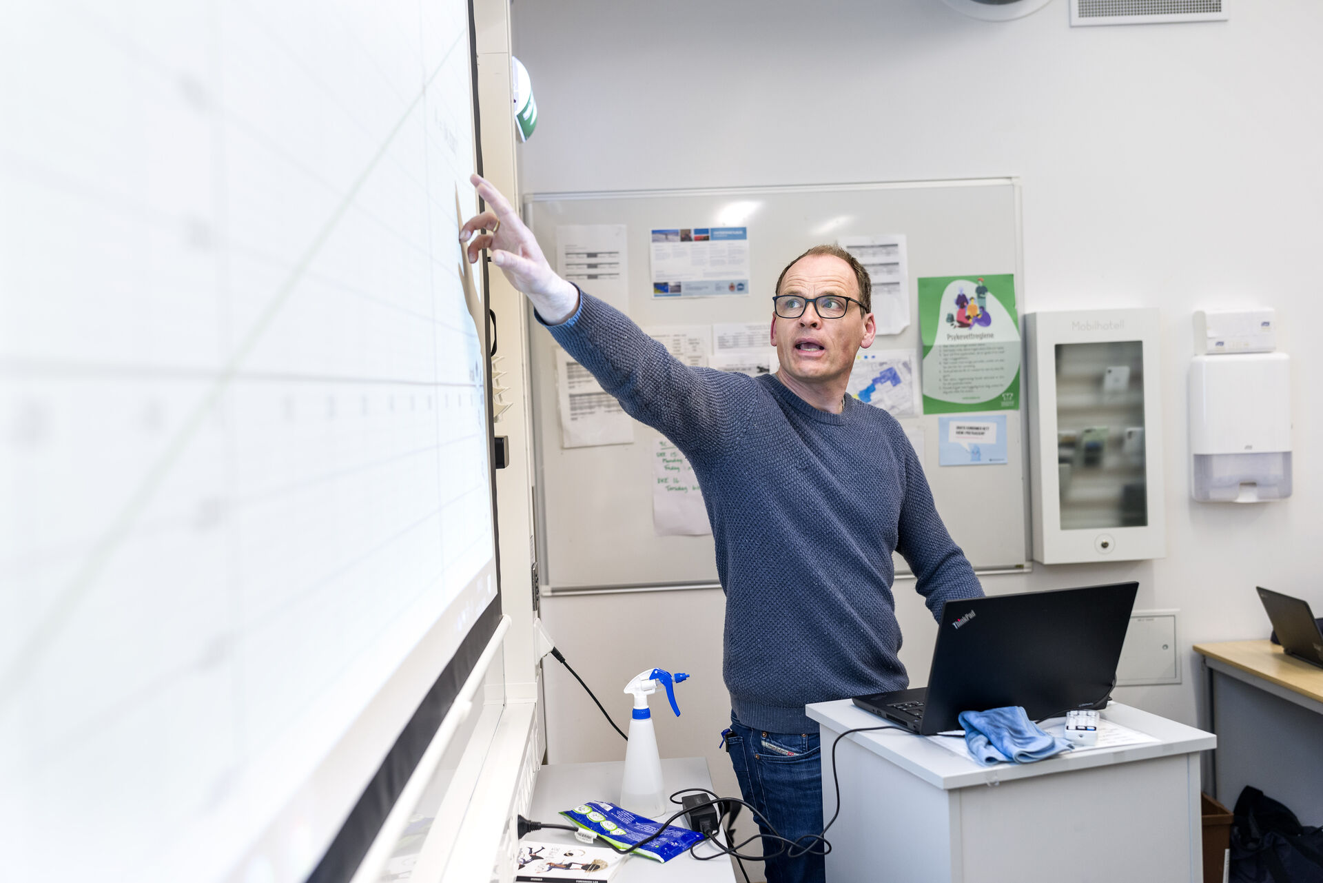 Mann med briller står og peker på en tavle i et klasserom