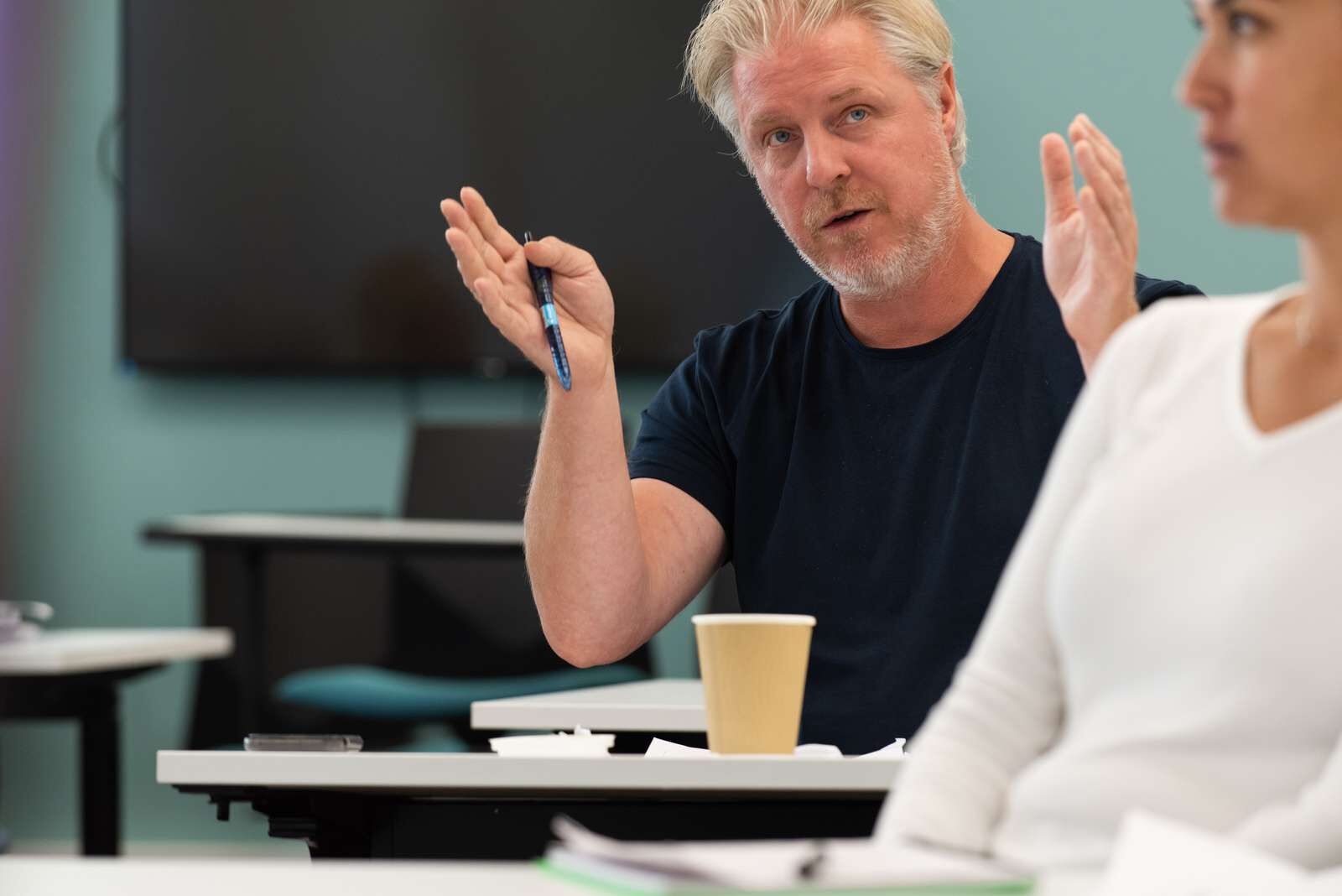 Dame står ved et bord og kommuniserer med 5 andre som sitter rundt bordet. Hun har front mot de og alle ser på henne.
