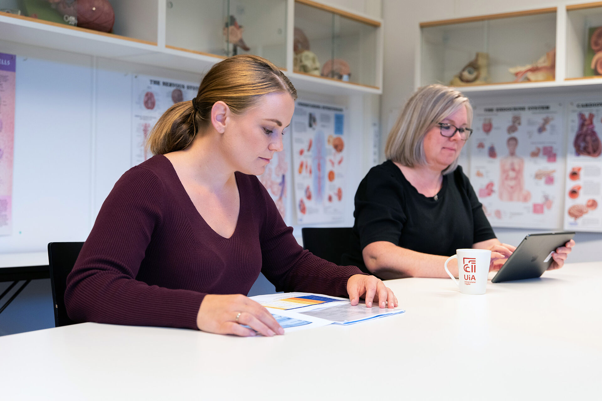 En kvinne med briller ser på en ipad og en annen kvinne ser i en brosjyre.