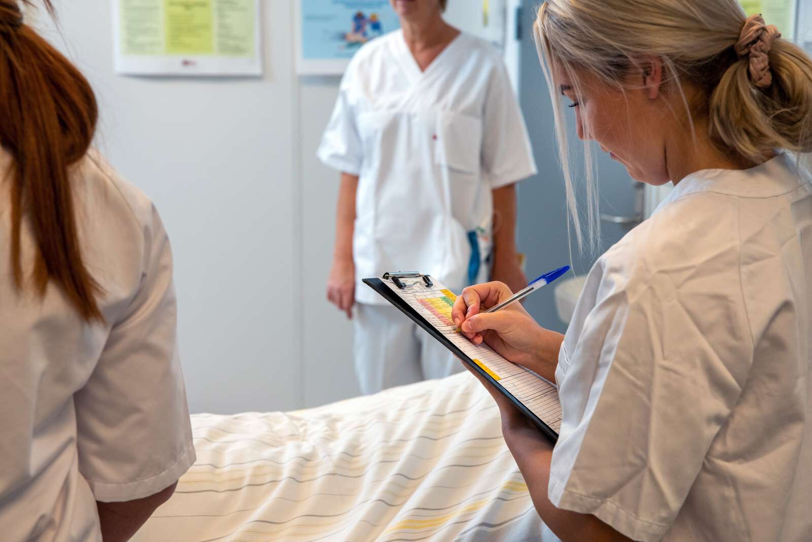 Kvinne i sykehusuniform står og noterer på en blokk ved siden av en sykehusseng