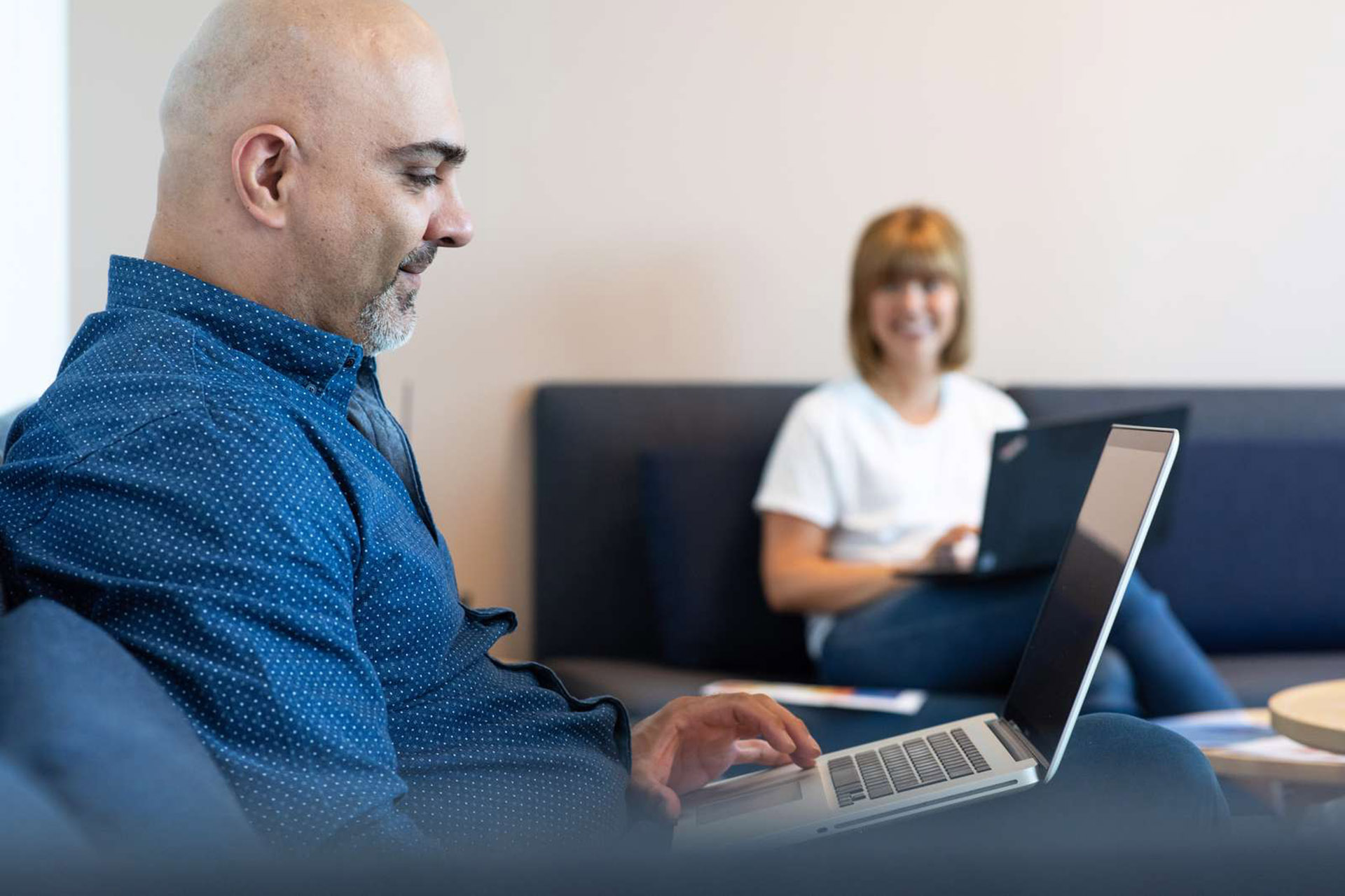 Mann uten hår sitter med laptop i fanget, mens kvinne i bakgrunnen ser på han