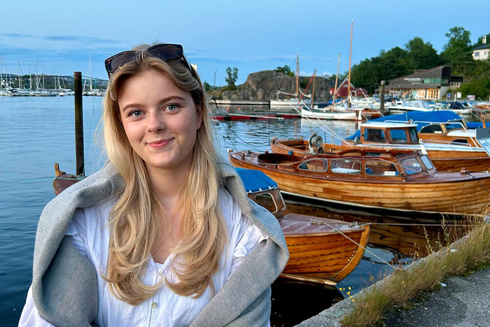 Bildet viser Mathilde med solbriller på hodet. Smiler. Båter og tresnekker i bakgrunnen.