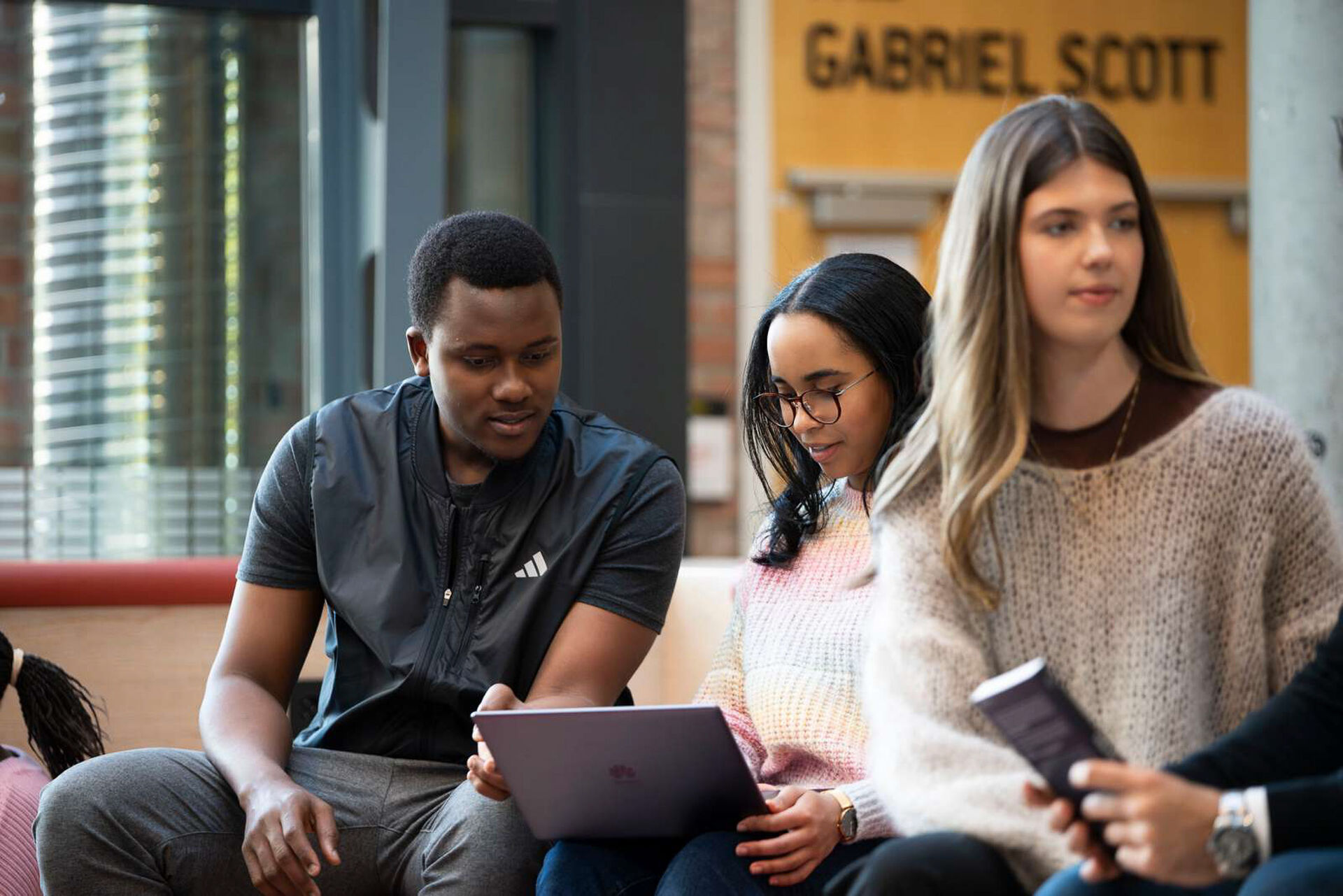 To studenter sitter og samarbeider over en laptop, mens en tredje samtaler med sidemannen.