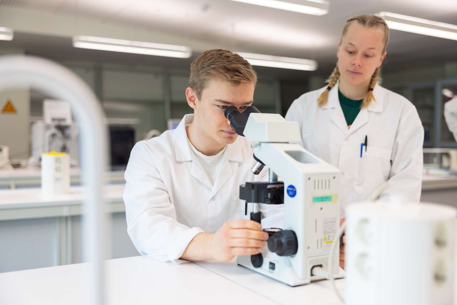 En student ser i mikroskop mens en annen student ser på