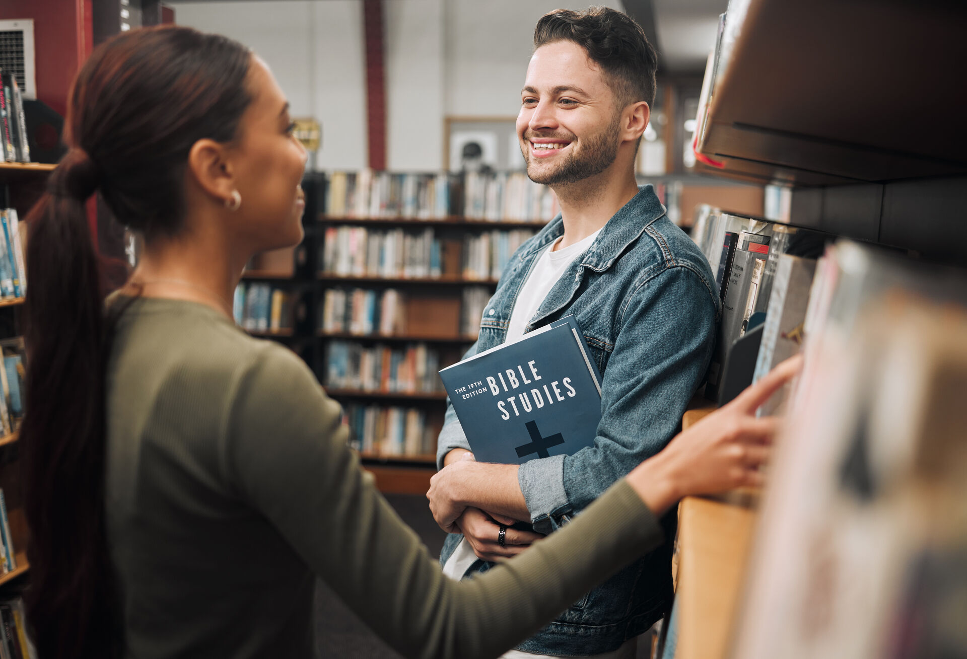 To studenter i biblioteket. Den ene ser i hylla etter bøker, mens den andre holder en religionsbok