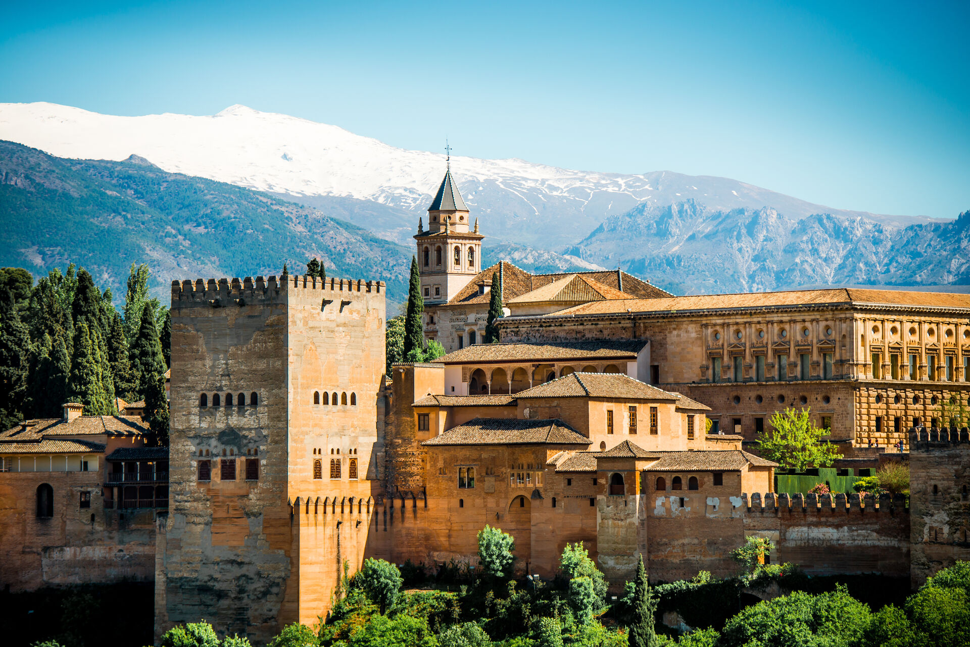 Alhambra i Granada. En stor bygning i gul kalkstein omringet av trær i forkanten av et fjell med snø på.