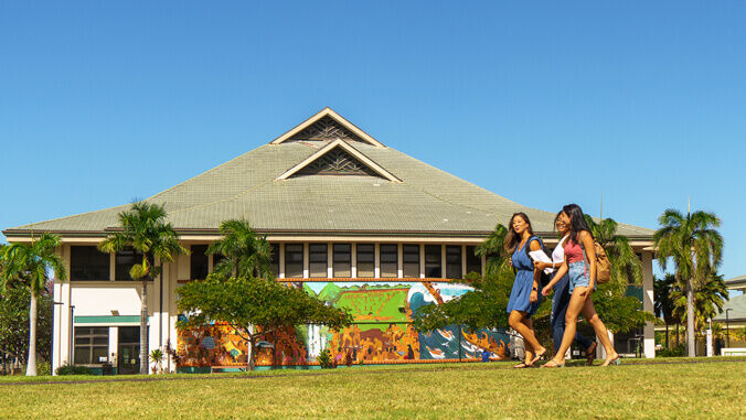 Photo of students crossing the lawn in front of one of the buildings on campus. The sun is shining and there are palm trees in the background