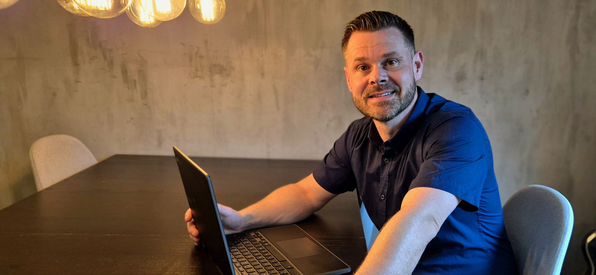 En mann sitter rundt et bord, med den ene hånden på pcen, mens han ser i kameraet
