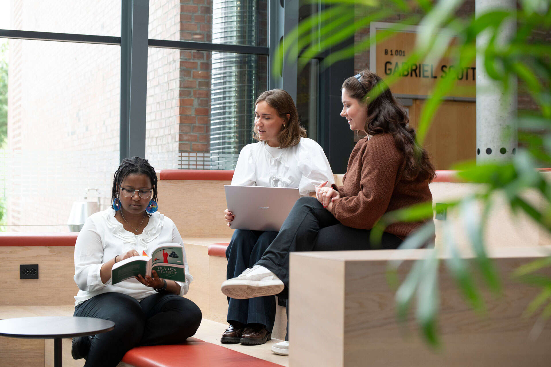 Three female students working together; one has a laptop on her lap and another is looking at a book